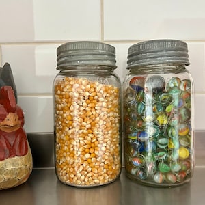 Peut inclure: Deux bocaux en verre avec des couvercles en métal, l'un rempli de grains de maïs soufflé jaunes et l'autre de billes colorées. Une figurine de coq en bois se trouve au premier plan.