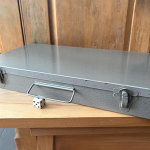 Peut inclure: Une boîte rectangulaire en métal gris avec une poignée et deux fermoirs. Un dé blanc avec des points noirs repose sur la surface en bois devant la boîte. La boîte est probablement utilisée pour le stockage.