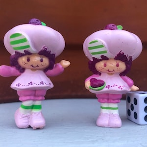 Puede incluir: Dos figuritas de Strawberry Shortcake de color morado y rosa con sombreros verdes y blancos. Una figurita sostiene un plato morado y verde.