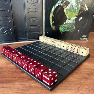 Peut inclure: Un jeu de société vintage intitulé "Duell" par Parker Brothers. Le jeu comprend un plateau de jeu à grille noire, des dés rouges et crème, et la boîte du jeu. La boîte présente des illustrations de deux personnages en capes. Le jeu est une stratégie de dés en tête-à-tête.