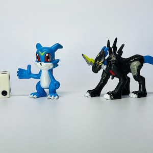 Peut inclure: Deux figurines d'action en plastique de personnages de dessins animés. La figurine bleue est une petite créature poilue avec une bande jaune sur le front et des yeux rouges. La figurine noire est une créature plus grande et plus menaçante avec une corne jaune et des yeux rouges. Les figurines sont posées comme si elles se faisaient face.