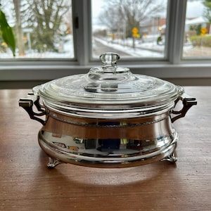 Op de afbeelding: Een verzilverde ovenschaal met een doorzichtig glazen deksel. De schaal heeft sierlijke handvatten en staat op drie kleine voetjes. Het deksel heeft een doorzichtige knop en een decoratief patroon. De schaal staat op een houten oppervlak.