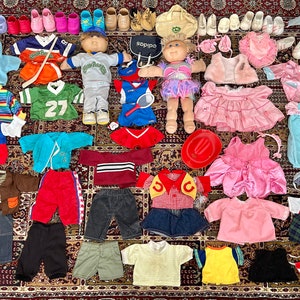 Könnte beinhalten: Eine Sammlung von Cabbage Patch Kids Puppen, die verschiedene Outfits tragen, darunter eine Baseballuniform, ein Cowboy-Outfit, ein Ballerina-Kostüm und eine Vielzahl anderer Kleidungsstücke. Die Puppen sind auf einem gemusterten Teppich mit verschiedenen Accessoires wie Schuhen, Hüten und Handtaschen angeordnet.