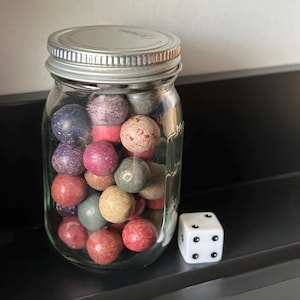 Puede incluir: Un frasco de vidrio transparente con tapa de metal plateado lleno de numerosas esferas pequeñas, redondas, texturizadas y multicolores en tonos rojo, azul, morado y beige. Un dado blanco de seis caras con puntos negros se encuentra junto al frasco.