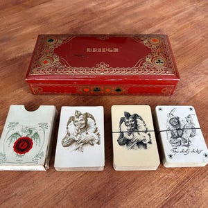 May include: A vintage red BRIDGE card box with gold decorative trim and a set of playing cards. The cards feature joker illustrations and the text "The jolly joker". The box and cards are on a wooden surface.