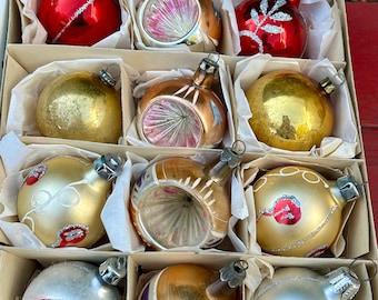 May include: A box of vintage Christmas ornaments with a variety of designs and colors, including gold, silver, red, and clear glass. The ornaments are in a white box with tissue paper.