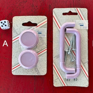 May include: Two packs of purple plastic cabinet knobs and handles. Pack A contains two round knobs. Pack B contains two L-shaped handles. Both packs have price tags.