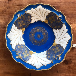 Puede incluir: Un plato decorativo azul con borde festoneado, con diseños de hojas blancas y detalles dorados. El centro y las áreas entre las hojas tienen intrincados patrones dorados. El plato está sobre una superficie de madera.
