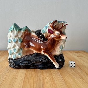Puede incluir: Figura de ciervo de cerámica con cuerpo marrón, manchas blancas y cornamenta. El ciervo salta sobre un tronco negro con un telón de fondo de árboles estilizados en tonos verdes y blancos. Hay un dado pequeño al lado de la figura.