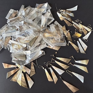 Peut inclure: Un grand tas de boucles d'oreilles emballées individuellement en coquille naturelle. Les boucles d'oreilles sont de formes et de tailles variées, certaines présentant une finition dorée. Les boucles d'oreilles sont présentées sur une surface noire.