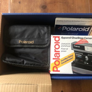 Peut inclure: Un appareil photo Polaroid OneStep noir dans sa boîte d'origine avec un étui de caméra noir. La boîte est rouge et blanche avec le texte "Polaroid" et "Appareil OneStep Camera".