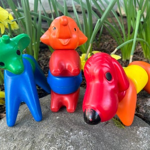 Può includere: Tre giocattoli impilabili di plastica colorati a forma di giraffa, elefante e cane. La girafa è verde e blu, l'elefante è arancione e rosso, e il cane è rosso e giallo.