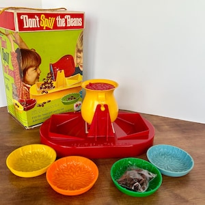 Puede incluir: Un juego vintage "Don't Spill the Beans" con una olla de frijoles roja, amarilla y naranja, una bandeja roja y cuatro cuencos de colores. La caja del juego presenta una ilustración colorida de niños jugando al juego.