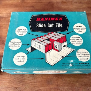 Peut inclure: Une boîte bleue et blanche pour ranger des diapositives 35 mm. La boîte est étiquetée "HANIMEX Slide Set File" et présente des illustrations du système de rangement des diapositives. La boîte peut contenir jusqu'à 240 diapositives en 12 groupes.