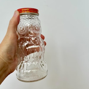 May include: Clear glass owl-shaped bottle with a red lid. The bottle has embossed details of an owl's face and body. The text "WISE PULLS" is visible on the bottom of the bottle. The bottle is held against a white background.