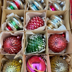Puede incluir: Una caja de adornos navideños vintage, que incluye un adorno de piña rojo y verde, un adorno plateado con una banda verde y roja, y un adorno de rayas rosa y blancas.