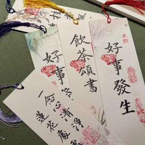 May include: A collection of decorative bookmarks with calligraphy and tassel accents. The bookmarks feature Chinese characters and red stamps, with tassels in blue, yellow, and red. The background has a green hue.