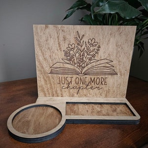 May include: Wooden book stand with a floral design above an open book, and the text "JUST ONE MORE chapter". The stand has a circular coaster and a rectangular slot. The wood has a natural finish.