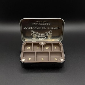 May include: A black pill organizer with a brown interior, divided into seven compartments labeled with the days of the week in gold lettering. The lid is open, revealing embossed text. The organizer is on a black background.