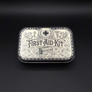 May include: A rectangular metal tin with a black and cream vintage-style design. The lid features the text "FIRST AID KIT" and "EMERGENCY SUPPLIES," along with illustrations of griffins, a medical cross, a key, a caduceus, and a small chest.