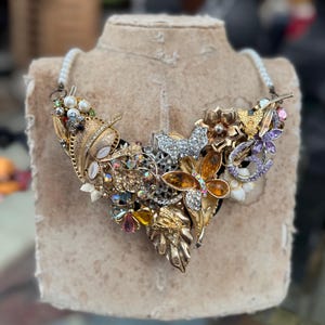 May include: A statement necklace with a heart-shaped pendant composed of various vintage jewelry pieces. The pendant features gold-toned leaves, butterflies, and floral elements, adorned with rhinestones and pearls. The necklace has a white beaded strand.