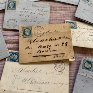 May include: A collection of antique envelopes in various shades of cream and light blue, scattered on a pink surface. The envelopes feature handwritten script, stamps, and postmarks. Some envelopes have the text "Bordeaux" and "Blanches".