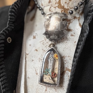 May include: A silver pendant necklace featuring a shield-shaped top piece and a rectangular, arched bottom piece. The bottom piece contains a small statue of a religious figure, flowers, and a piece of paper. The necklace is on a black beaded chain.