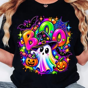 Puede incluir: Una camiseta negra con un gráfico vibrante y colorido de un fantasma con sombrero de bruja, rodeado por la palabra BOO en letras de neón, una calabaza, un cubo de dulces, murciélagos, telarañas y varios dulces sobre un fondo.