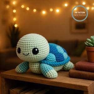 Puede incluir: Una tortuga de ganchillo hecha a mano en tonos verdes y azules, con una cara sonriente y grandes ojos negros. La tortuga está sobre una superficie de madera. La imagen incluye una etiqueta de descarga digital.