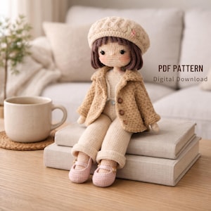 Op de afbeelding: Een gehaakte pop met een beige jas, broek en baret, met roze schoenen. De pop heeft bruin haar en roze wangen. De pop zit op twee boeken. De tekst "PDF PATTERN Digital Download" is zichtbaar.