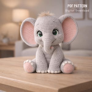 Op de afbeelding: Een gehaakte grijze olifant met grote roze oren, grote groene ogen en een kleine slurf. De olifant zit met uitgestrekte poten en heeft roze accenten op de voeten. De tekst "PDF PATTERN Digital Download" staat rechtsboven.