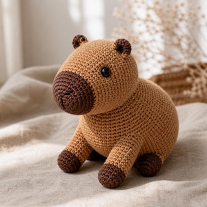 Op de afbeelding: Een handgemaakte gehaakte capybara knuffel. De knuffel is voornamelijk lichtbruin met donkerbruine accenten op de neus, oren en voeten. Het heeft zwarte veiligheidsoogjes en zit op een neutraal gekleurde stof.