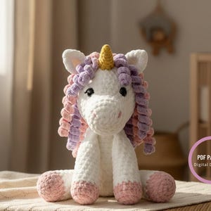 Peut inclure: Une licorne blanche faite à la main au crochet, avec des cheveux bouclés roses et violets, une corne jaune et des sabots roses. L'animal en peluche est assis sur une couverture crème.