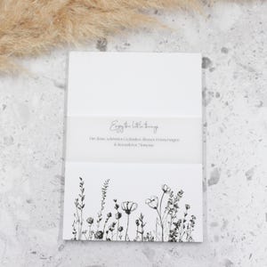 Könnte beinhalten: Weiße Karte mit einer schwarzen Tintenzeichnung von Wildblumen und dem Text "Enjoy the little things" und "For your beautiful celebrations, little memories & besonderen moments".