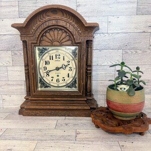 May include: A wooden mantel clock with an arched top, a cream-colored face, black Arabic numerals, and ornate corner details. Beside it sits a small green succulent in a round, multi-colored ceramic pot resting on a carved wooden base.