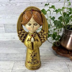 May include: A gold angel figurine with red gem accents. The angel has brown hair, a white face, and is wearing a gold robe. The angel is holding its hands together in prayer.