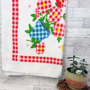 May include: White kitchen towel with a red gingham border and floral design. The towel features a blue gingham vase overflowing with red gingham flowers with yellow centers. Small yellow chicks and pink dots are scattered around the flowers.