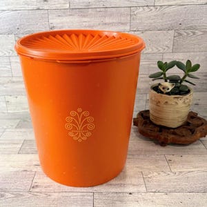 May include: A bright orange cylindrical plastic canister with a matching lid featuring a sunburst pattern. The front of the container displays a small, intricate yellow floral-inspired graphic design centered on the smooth, solid orange surface.