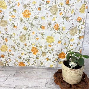 Könnte beinhalten: Weißer Stoff mit Blumenmuster mit gelben, orangefarbenen und beigefarbenen Rosen und grünen Ranken. Eine kleine Topfpflanze mit einer Katzenfigur steht auf einem Holztablett im Vordergrund.