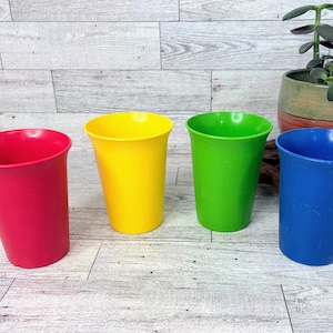 Puede incluir: Un juego de cuatro vasos de plástico de colores sólidos en rojo, amarillo, verde y azul. Cada vaso presenta una forma cilíndrica lisa y cónica con un borde ancho y una base plana, colocados en posición vertical en una fila.