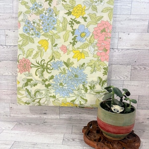 May include: A rectangular piece of fabric featuring a floral pattern with yellow, light blue, and pink flowers on a cream background. A small green and red ceramic planter with a succulent sits on a carved wooden base nearby.