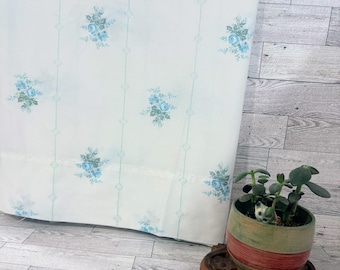 Op de afbeelding: Een wit stoffen gordijn met een herhalend patroon van lichtblauwe bloemenboeketten en dunne verticale groenblauwe lijnen. Naast het gordijn staat een ronde keramische plantenpot met horizontale rode en groene banden met een kleine vetplant.