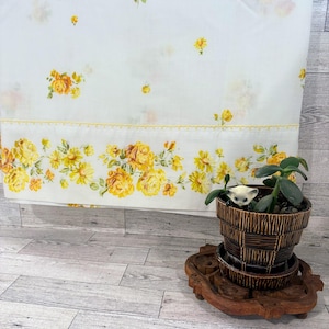 May include: A white fabric panel featuring a yellow floral print with scattered small roses and a dense border of yellow roses, accented by a yellow decorative trim. A brown textured ceramic planter holds a green plant and a small cat figurine.