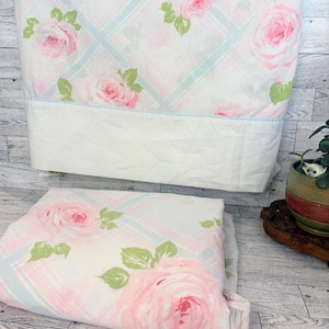 May include: White bed sheets with a pink rose floral pattern and light blue plaid design. The sheets are folded and displayed, showcasing the detailed rose and leaf design. The bedding set is made of a soft material.