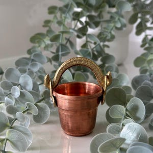 May include: A small, copper-colored bucket with a gold-colored handle and accents. The bucket has a cylindrical shape and is surrounded by faux greenery. The handle is arched and textured, adding a decorative element to the piece.