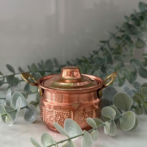 Peut inclure: Une petite marmite en cuivre avec un couvercle et deux poignées. La marmite présente un motif floral en relief sur la base et une finition brillante. Les poignées et le dessus du couvercle sont de couleur dorée.