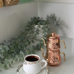 以下が含まれることがあります： 金色の縁取りが付いた白いコーヒーカップが、濃いコーヒーが入ったお揃いのソーサーの上に置かれています。装飾的なハンドルと蓋が付いた銅製のティーポットがカップの隣にあります。セットは白い表面にあります。