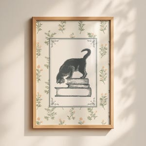 May include: A framed wall art print featuring a black cat standing on a stack of three books. The central illustration is enclosed in a decorative border, set against a cream background patterned with small green sprigs and tiny orange flowers.