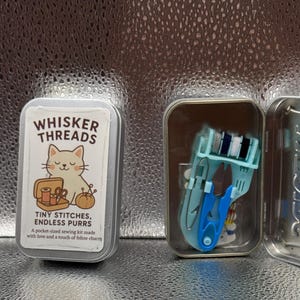 May include: A small, rectangular tin with the text "WHISKER THREADS" and a cat illustration. The tin contains a pocket-sized sewing kit with blue scissors and thread. The text "TINY STITCHES, ENDLESS PURRS" is also visible.