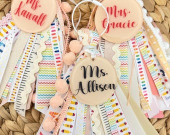 May include: Three personalized teacher gifts with ribbons and pom-poms. Each features a round, peach-colored acrylic tag with a teacher's name in elegant script: "Ms. Kawall", "Mrs. Gracie", and "Ms. Allison". The ribbons include pencil and ruler patterns.